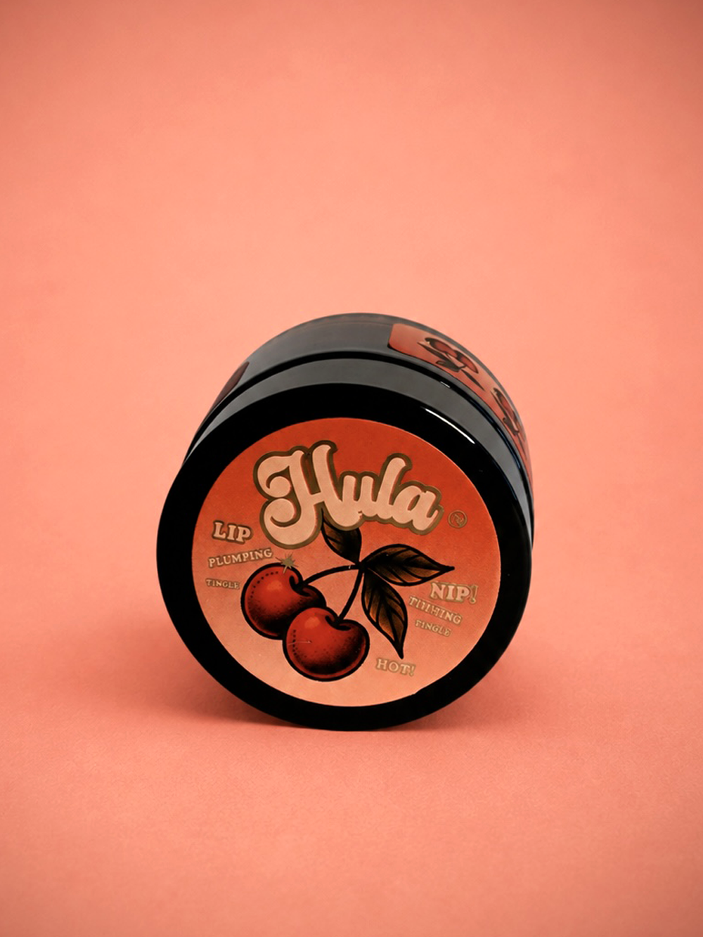 A jar of Hula Cherries Jubilee Lip and Nip Balm Pot with a black container and a round orange label featuring a cherry illustration, set against a solid peach-pink background.