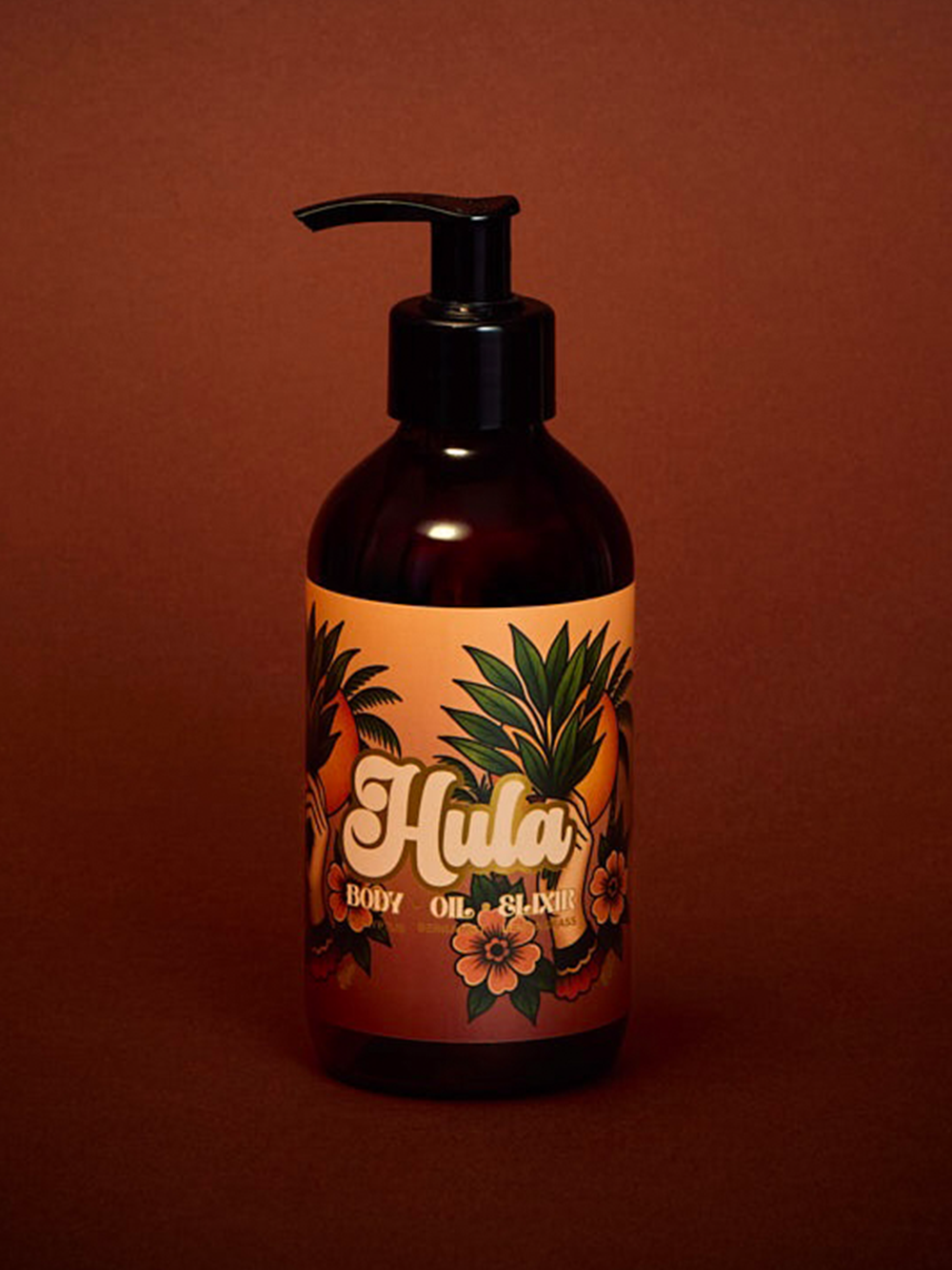 An amber glass pump bottle of Hula Gold Body Oil with a black pump, set against a solid dark reddish-brown background. The label features a warm orange background with illustrations of green palm leaves and pink hibiscus flowers, with the brand name 'Hula' written in a stylised white font.