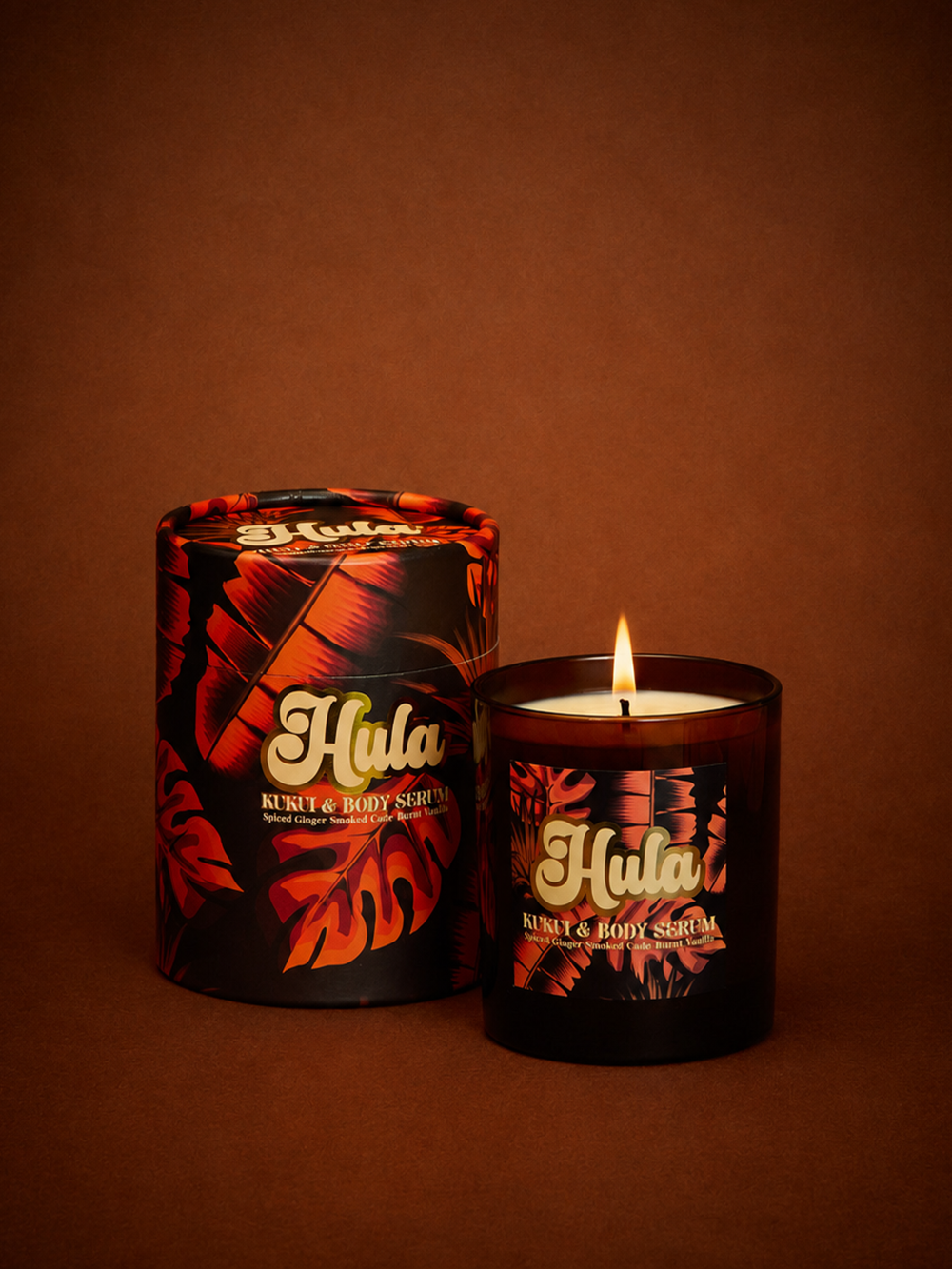 A lit Hula Nights candle in a dark amber glass jar stands next to its matching cylindrical packaging box, both decorated with a black tropical leaf pattern in orange and red tones, set against a solid dark brown background.