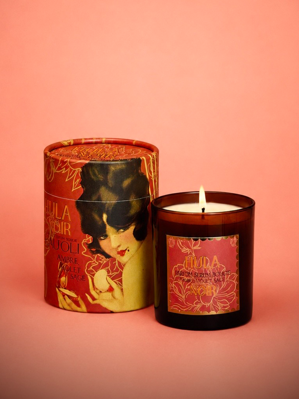 An amber glass jar candle next to its matching cylindrical packaging box, both featuring a vintage Art Deco-style illustration of a woman and the brand name 'Hula Noir'. The product is set against a solid coral-pink background.