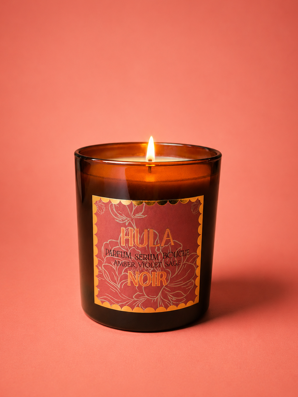 An amber glass jar candle with a lit wick, featuring a red and gold label that reads 'HULA NOIR' on a solid coral-pink background.