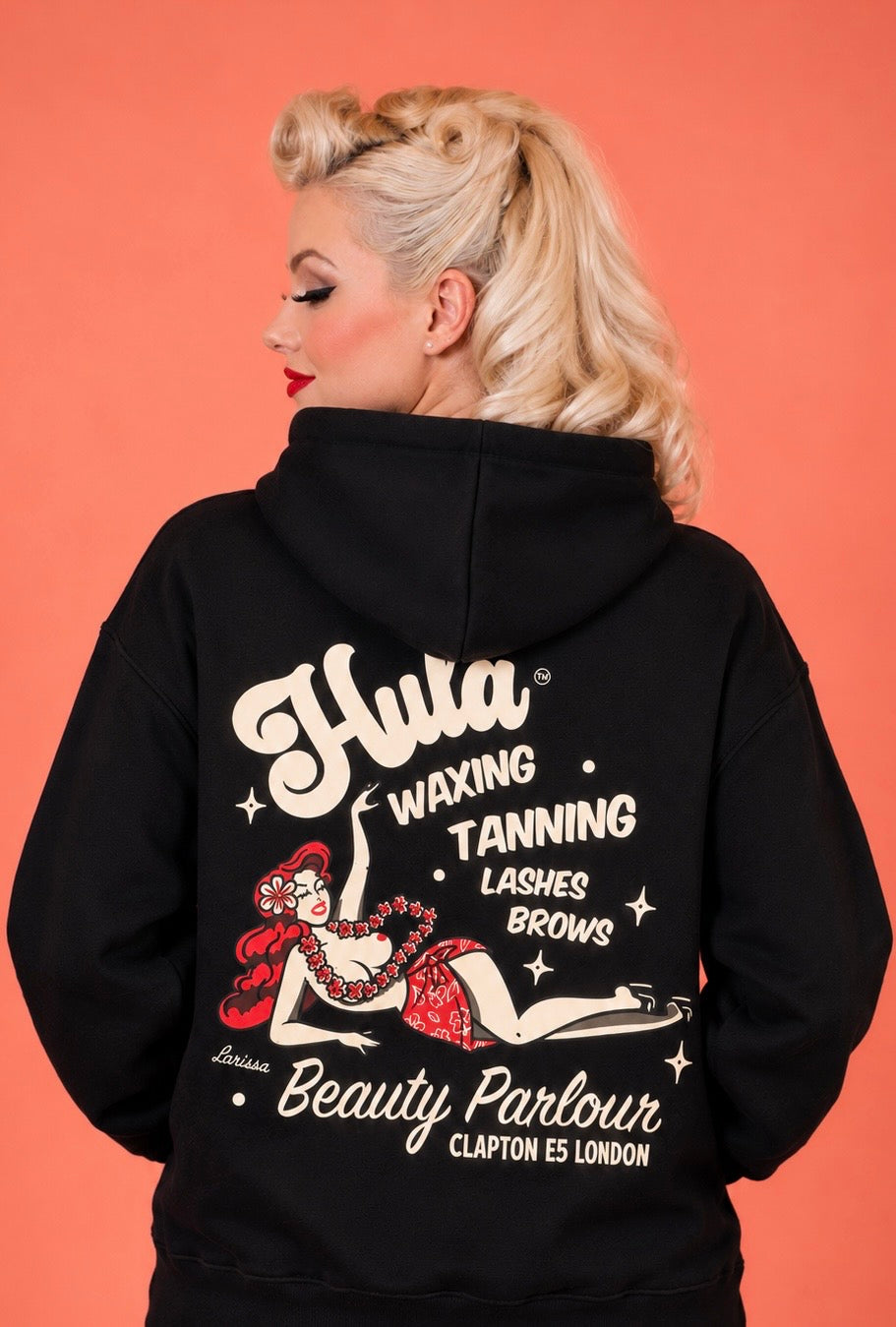 A woman with blonde hair is shown from the back wearing a black hoodie with a large vintage-style graphic on the back. The graphic features cream text reading 'Hula Waxing Tanning Lashes Brows Beauty Parlour' and a red-and-white pin-up illustration, against a solid coral-pink background.