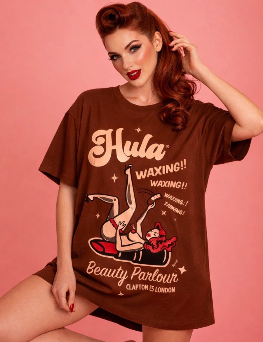 A woman modeling a brown graphic t-shirt with retro pin-up art and text reading 'Hula Waxing!! Beauty Parlour Clapton ES5 London' against a solid pink background.