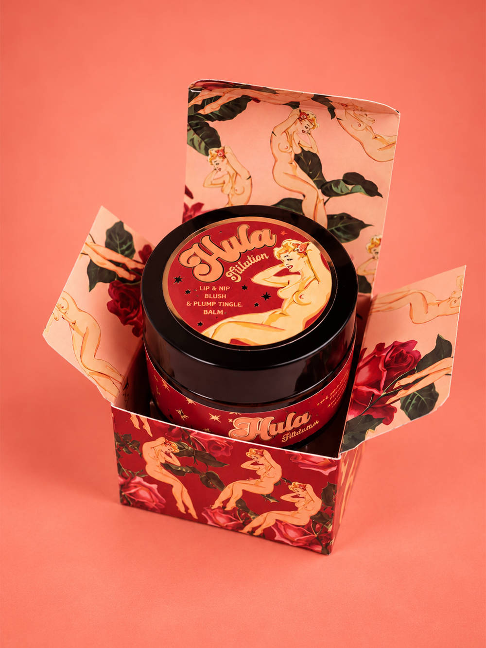 A jar of Hula Titilation™ Lip & Nip Balm Pot sits open in its illustrated red and pink box against a solid coral background. The packaging features retro pin-up woman illustrations and the brand name 'Mula' in stylised lettering.