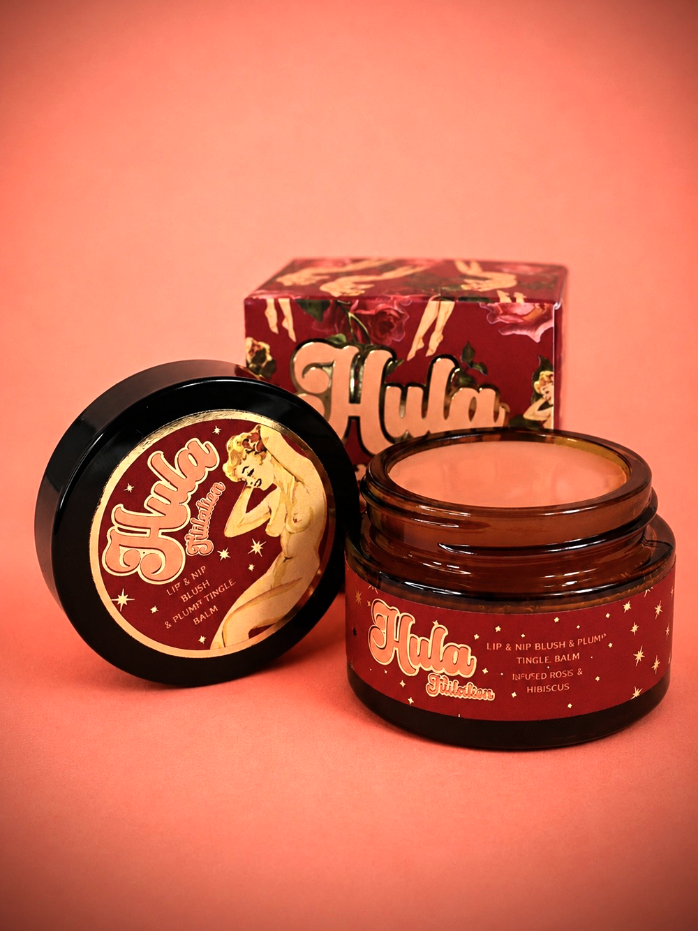 An open amber glass jar of Titilation™ Lip & Nip Balm and its matching lid with a decorative label, placed in front of a branded red box on a peach-pink studio background. The product appears to be a lip and eye blush & peach tingle balm from Hula Beauty.