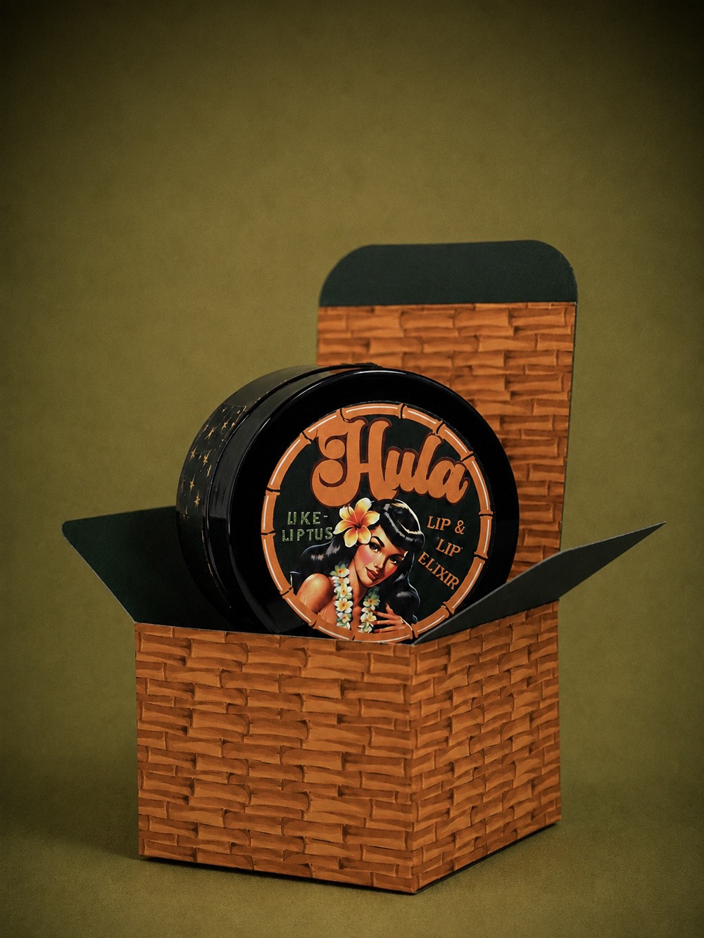 A round tin of Hula Uke-Lyptus Lip and Nip Balm displayed inside a decorative brown woven box on a dark olive background. The tin is black with orange lettering and an illustration of a hula dancer.
