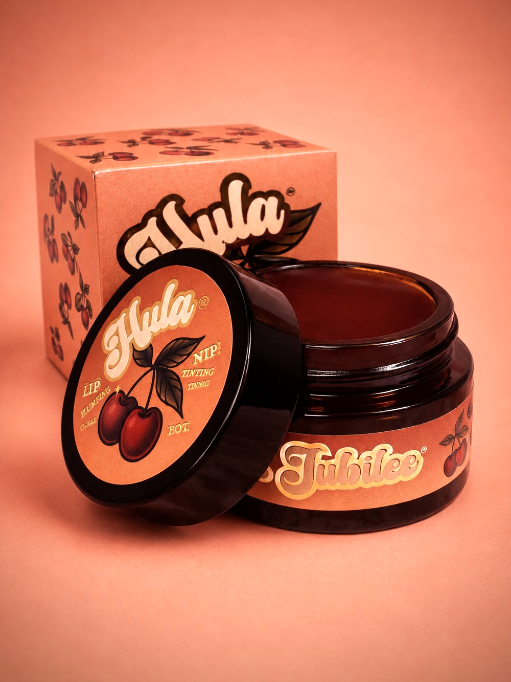 An open amber glass jar of Cherries Jubilee™ Lip & Nip Balm with its lid leaning in front, next to its branded cardboard box, on a solid peach-pink background.