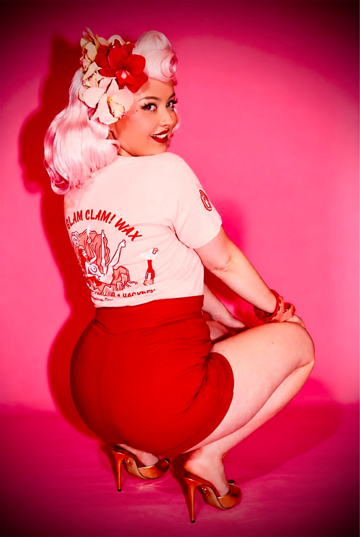 A woman in a retro pin-up pose wearing a light pink graphic t-shirt and a red skirt against a vibrant pink background.