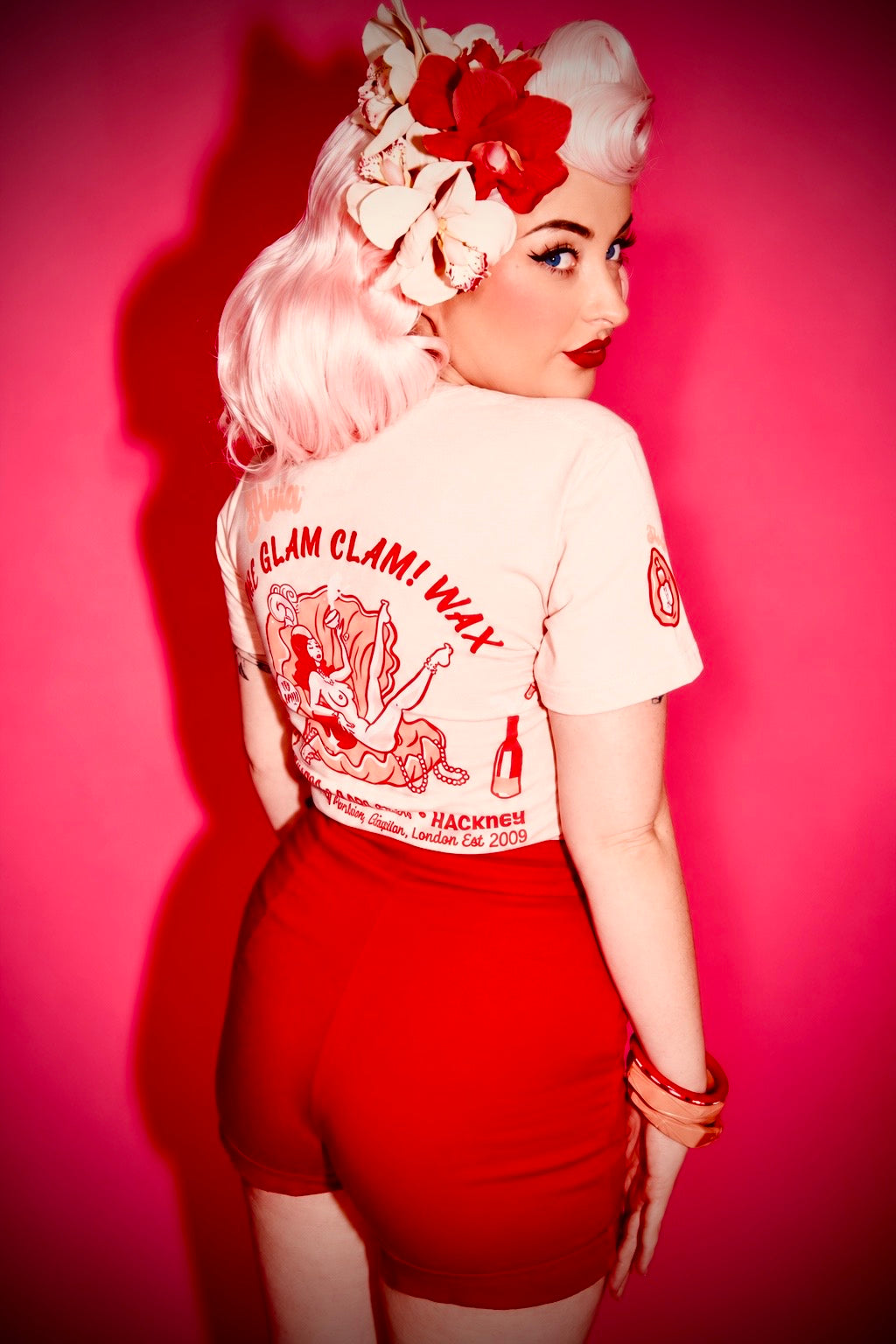 A woman posing from behind wearing a cream graphic t-shirt with red text that reads 'GLAM CLAM! WAX', paired with red shorts, set against a vibrant pink background.