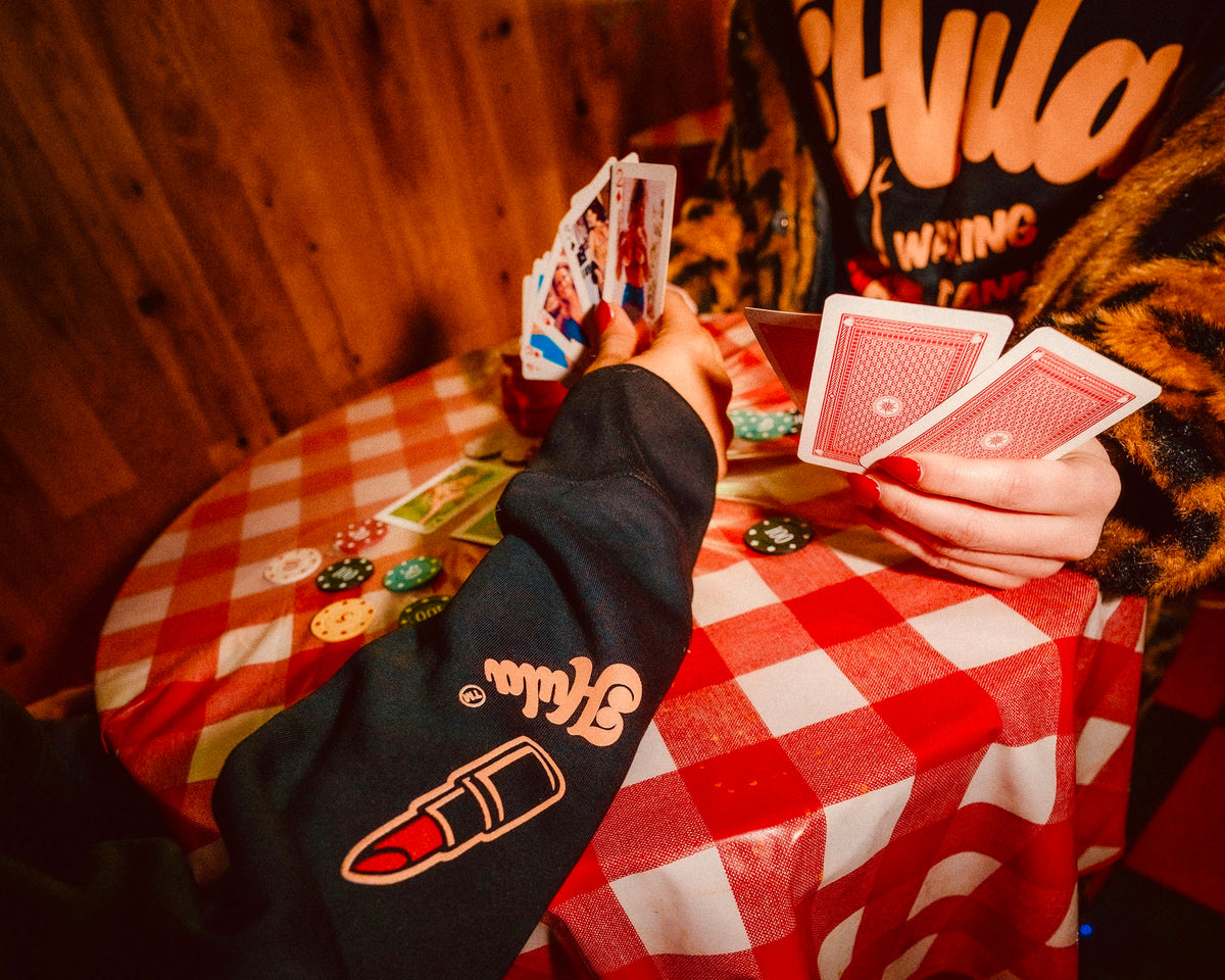 A person seated at a round table with a red-and-white checkered tablecloth, holding playing cards. The person wears a black sweatshirt with a large pink lipstick graphic and orange lettering on the sleeve, along with a leopard-print jacket.