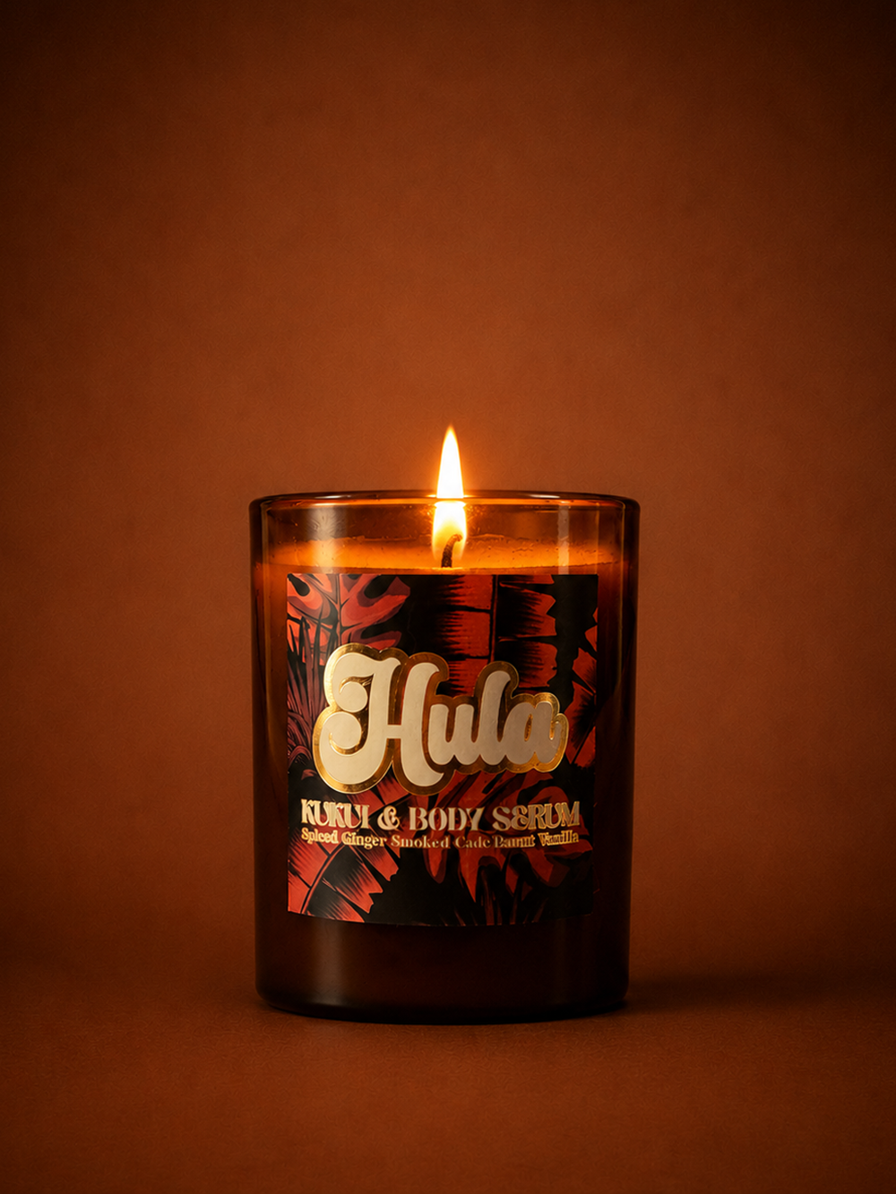 A lit candle in a dark amber glass jar with a red tropical leaf label featuring the brand name 'Hula' in gold script, set against a warm brown background.