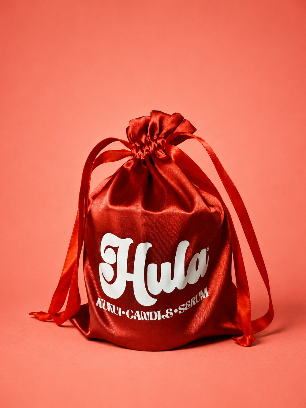 A red satin drawstring pouch with white 'Hula' branding and smaller 'KUKUI-CANDLE SERUM' text, photographed on a solid coral-pink background.