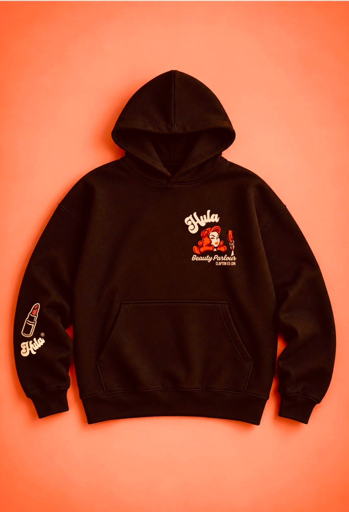 A black pullover hoodie laid flat against a solid orange background, featuring a red and white graphic on the left chest with the text 'Hula Beauty Parlour' and a lipstick on the right sleeve.