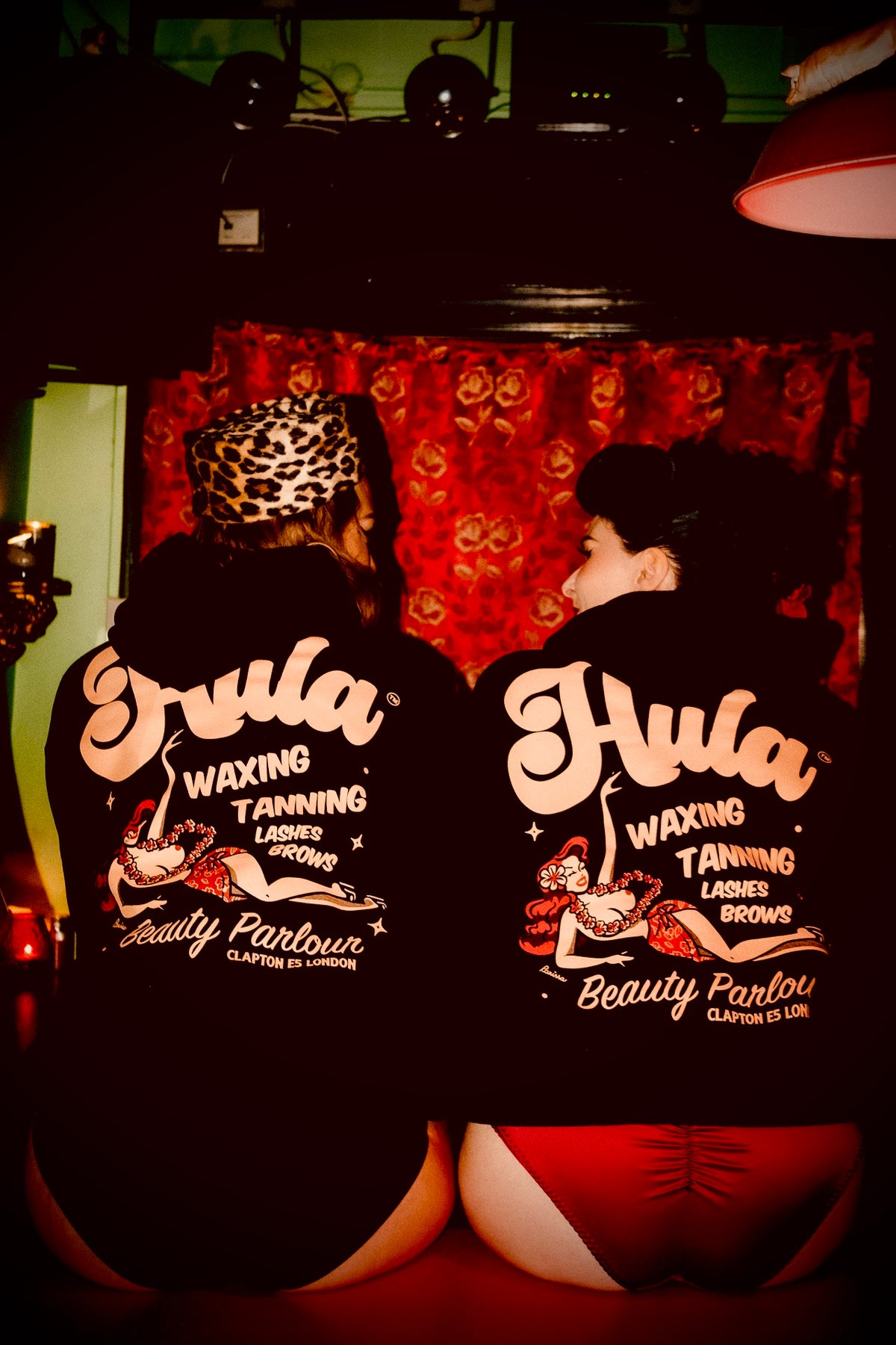 Two women seated with their backs to the camera, wearing black hoodies with large 'Hula Waxing Tanning Beauty Parlour' graphics on the back. The scene is dimly lit with red ambient lighting, and a red patterned curtain hangs behind them.