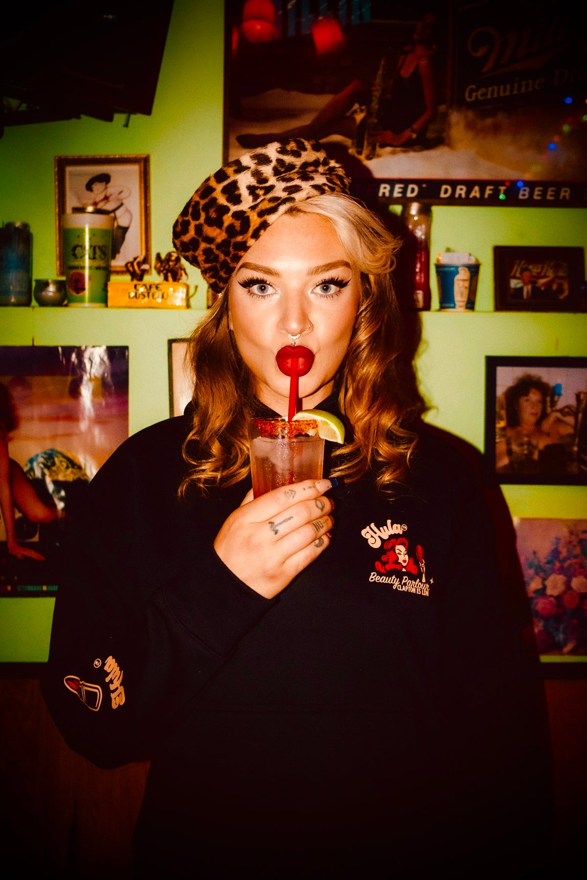 A woman wearing a leopard-print beret and a black hoodie with a red and white graphic on the chest, holding a drink with a red straw in front of a green wall with framed posters.