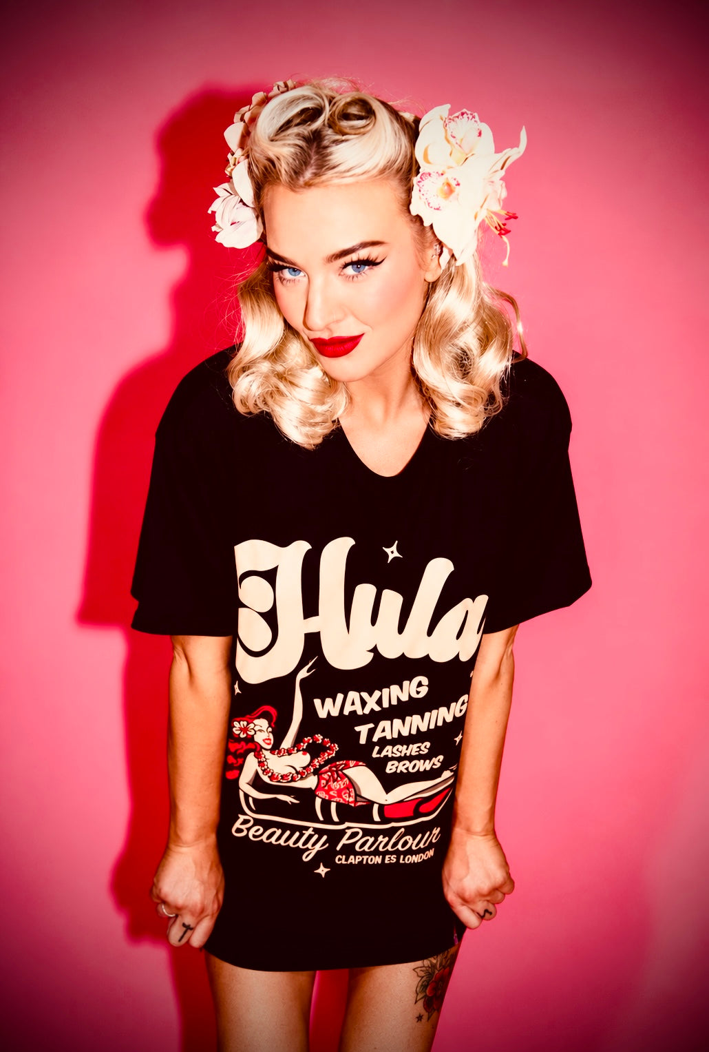 A woman with vintage blonde curls, white flowers in her hair, and red lipstick wears a black graphic t-shirt with retro-style cream lettering that reads 'Hula Waxing Tanning Beauty Parlour'. She is posed against a solid pink background.