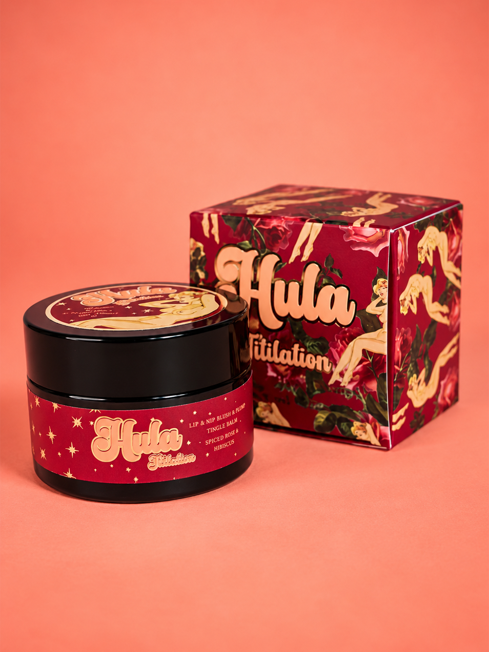 A black cosmetic jar of Titilation™ Lip & Nip Balm Pot and its matching decorative box from the brand Hula Beauty,  featuring a red and gold tropical print, set against a solid coral background.