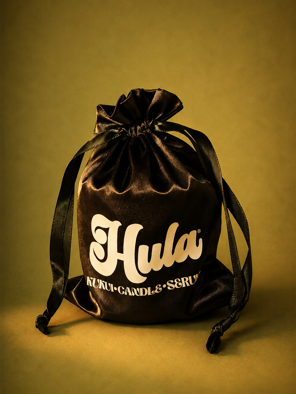 A black velvet drawstring pouch with 'Hula Kuki Candle & Scrum' printed in cream, set against a warm olive-yellow background.
