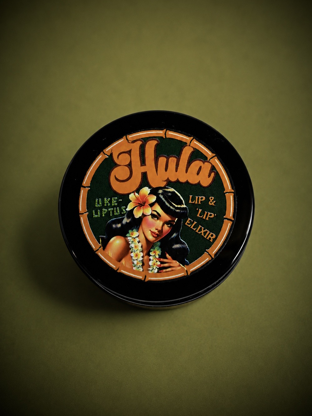 A round black tin of Hula Uke-Lyptus Lip & Nip Balm with a retro Hawaiian-themed illustration on the lid, set against a dark olive green background.