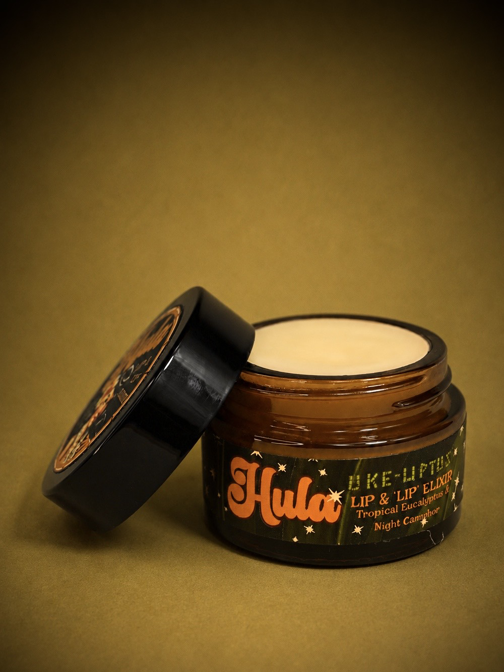 An open amber glass jar of Hula Uke-Lyptus Lip and Nip Balm with its black lid leaning against it, showing the cream-colored tint inside. The product is set against a solid, warm olive-brown background.